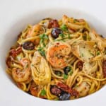 A close-up view of a large white pasta bowl filled with Mediterranean past with shrimp garnished with chopped Italian parsley.