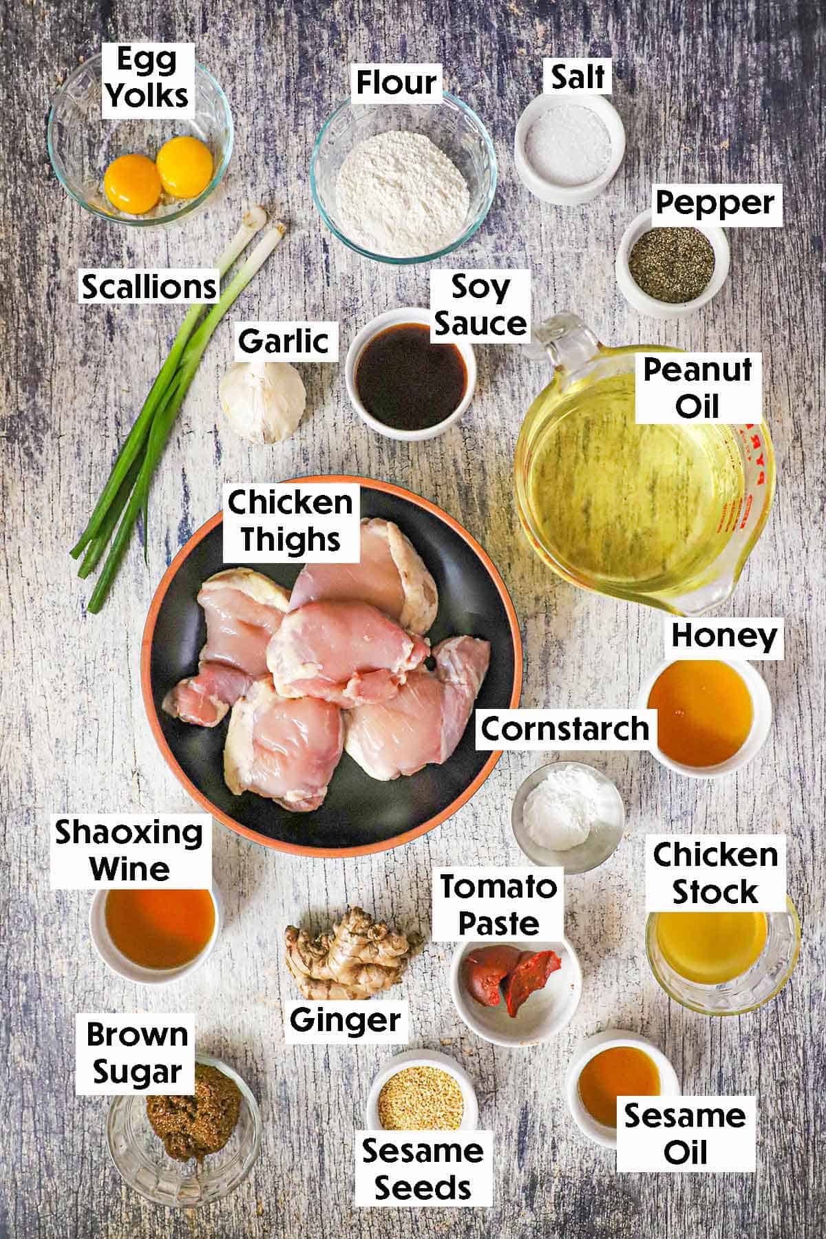 An arrangement of ingredients for homemade sesame chicken on a grey wooden background including chicken thighs, soy sauce, oil, egg yolks, honey, garlic, scallions, ginger, chicken stock, seasonings.