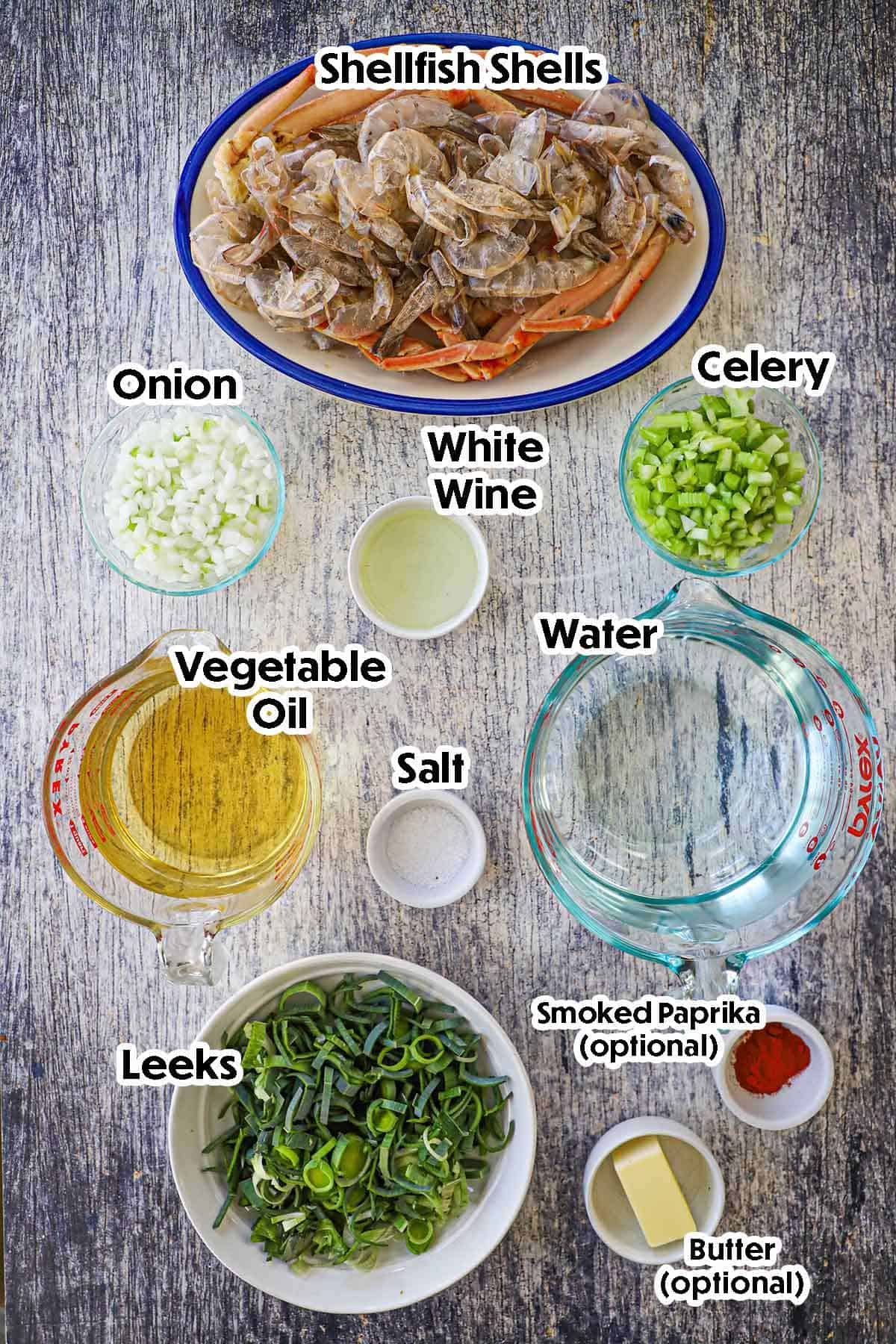 An arrangement of ingredients for homemade shellfish stock on a grey wooden background including shellfish shells, onion, celery, wine, leeks, oil, and seasonings.