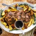 An overhead view of a large white oval platter that is filled with a sliced slow-roasted lamb shoulder with crispy potatoes and a small bowl of red wine au jus.