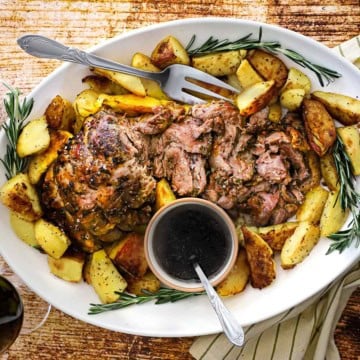 An overhead view of a large white oval platter that is filled with a sliced slow-roasted lamb shoulder with crispy potatoes and a small bowl of red wine au jus.