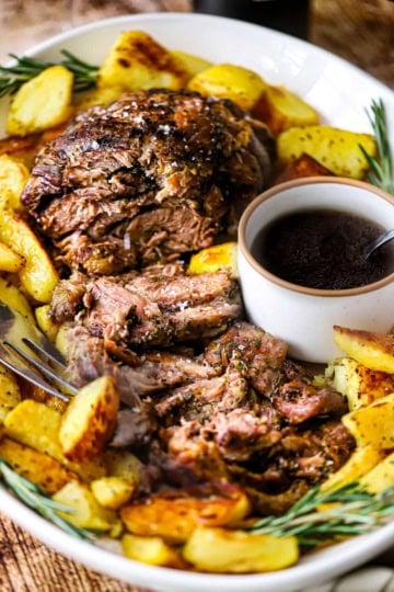 A large white oval platter filled with a slow-roasted lamb shoulder that has been roughly slices and is surrounded by crispy potato wedges and a small bowl filled with a red wine au jus.