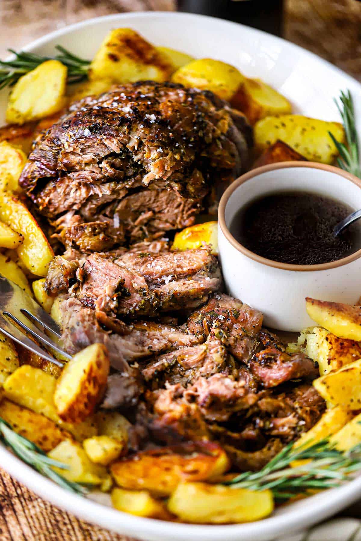 A large white oval platter filled with a slow-roasted lamb shoulder that has been roughly slices and is surrounded by crispy potato wedges and a small bowl filled with a red wine au jus.