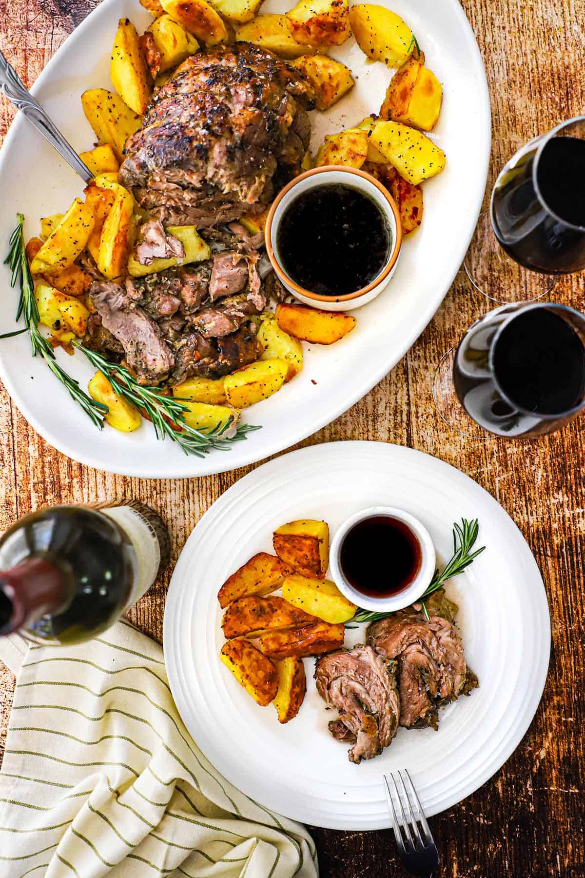 An overhead view of a large platter filled with sliced slow-roasted lamb shoulder with crispy potatoes next to a dinner plate filled with a serving of the same and two glasses of red wine nearby.