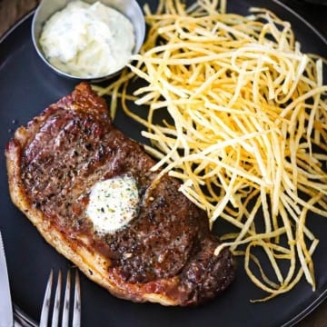 An overhead view of French Bistro-Style Steak Frites with a reverse-seared New York Strip steak on a plate next to a pile of shoestring French fries and a small vessel of garlic basil aioli.