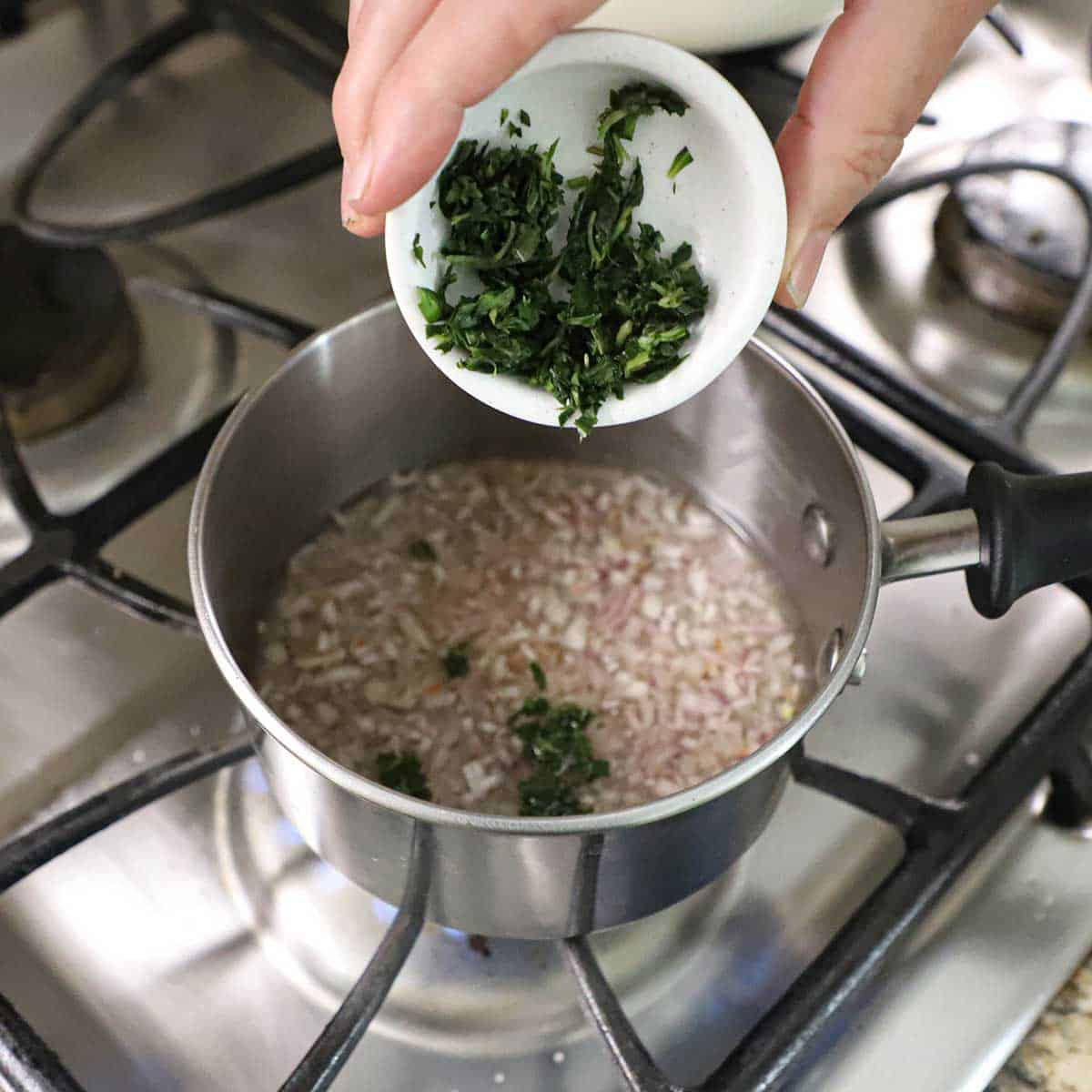 A person dumping chopped, fresh tarragon from a small white bowl into a small saucepan containing simmering vinegar, white wine, and chopped shallots.