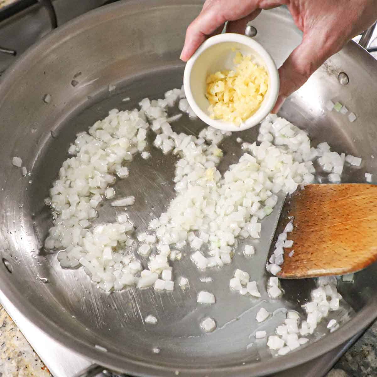 A person dumping minced garlic from a small bowl into a large silver skillet that contains chopped onion that is being sautéed in oil.