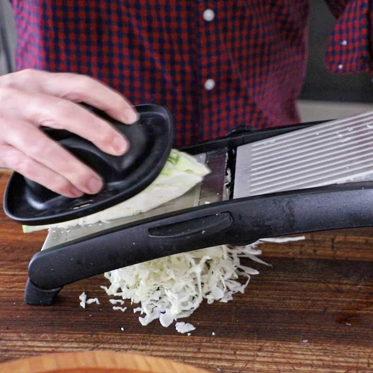 A person using a mandolin to finely shred green cabbage on a wooden cutting board.