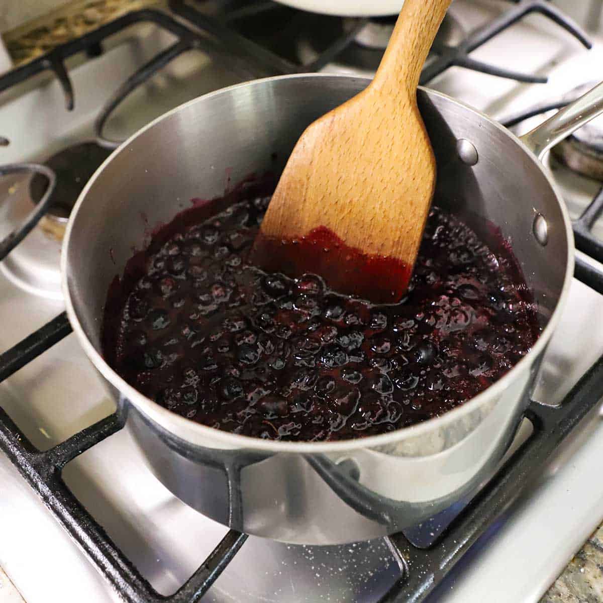 A person using a wooden spatula to stir simmering blueberries in a silver saucepan on a gas stove.