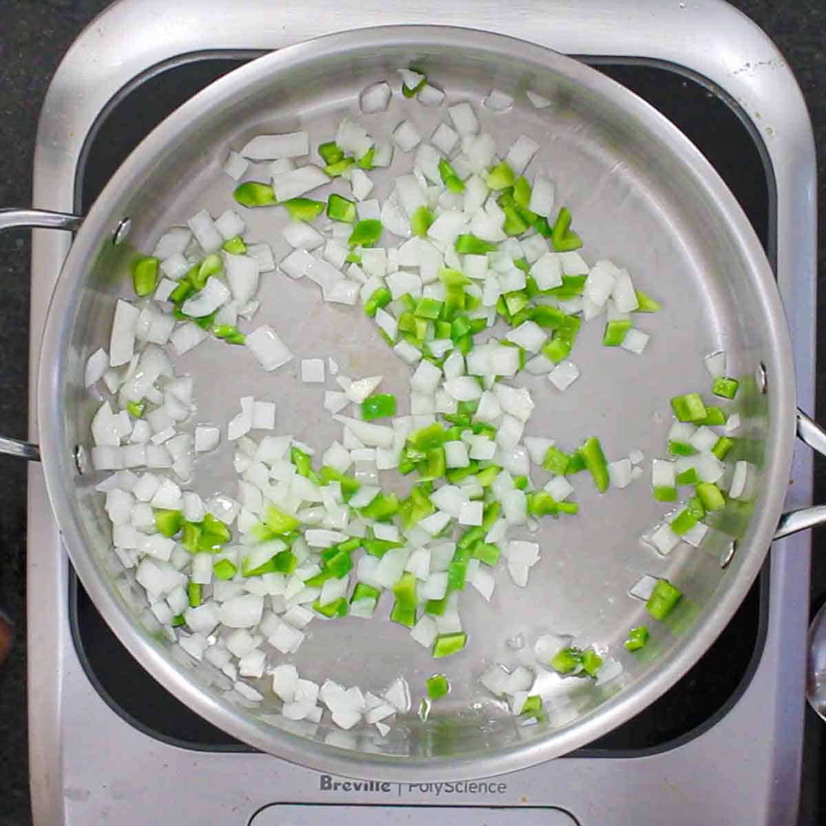 An overhead view of chopped onion and green bell pepper being sautéed in oil in a large stainless steel skillet.