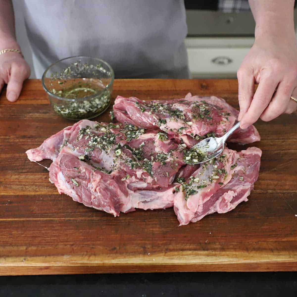 A person using a spoon to spread a fresh herb and garlic paste over the top of an uncooked lamb shoulder on a wooden cutting board.