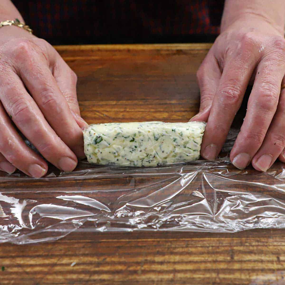 A person rolling softened compound parsley and garlic butter into a log in plastic wrap on a wooden cutting board.