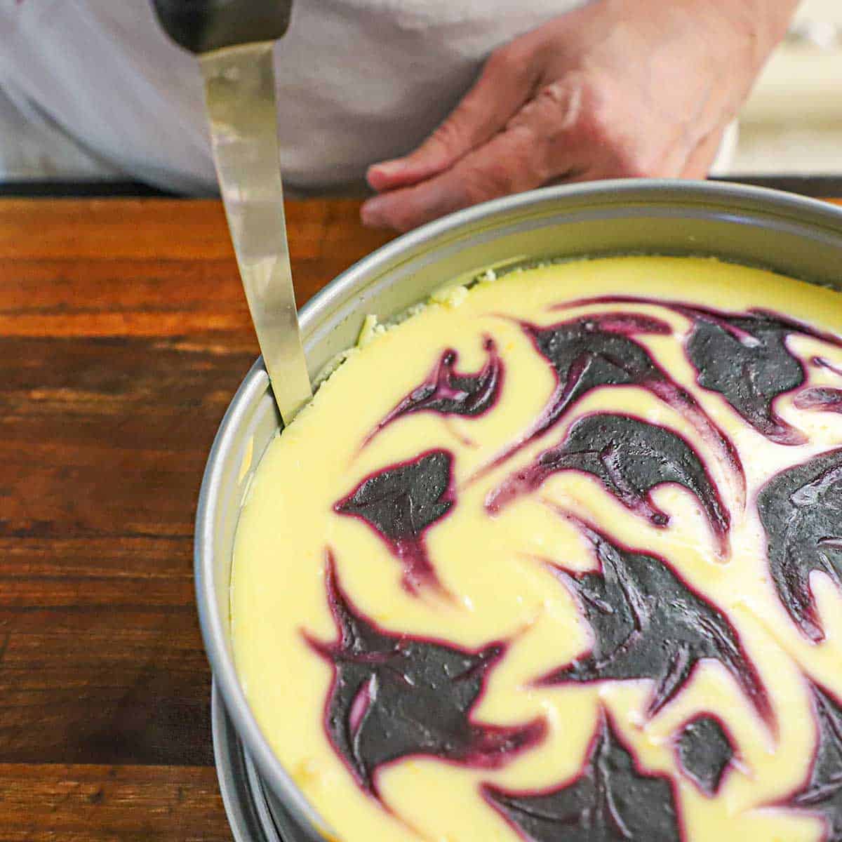 A person using a knife to cut the edges of a cooked lemon blueberry cheesecake from the side of the springform pan that is what baked in.