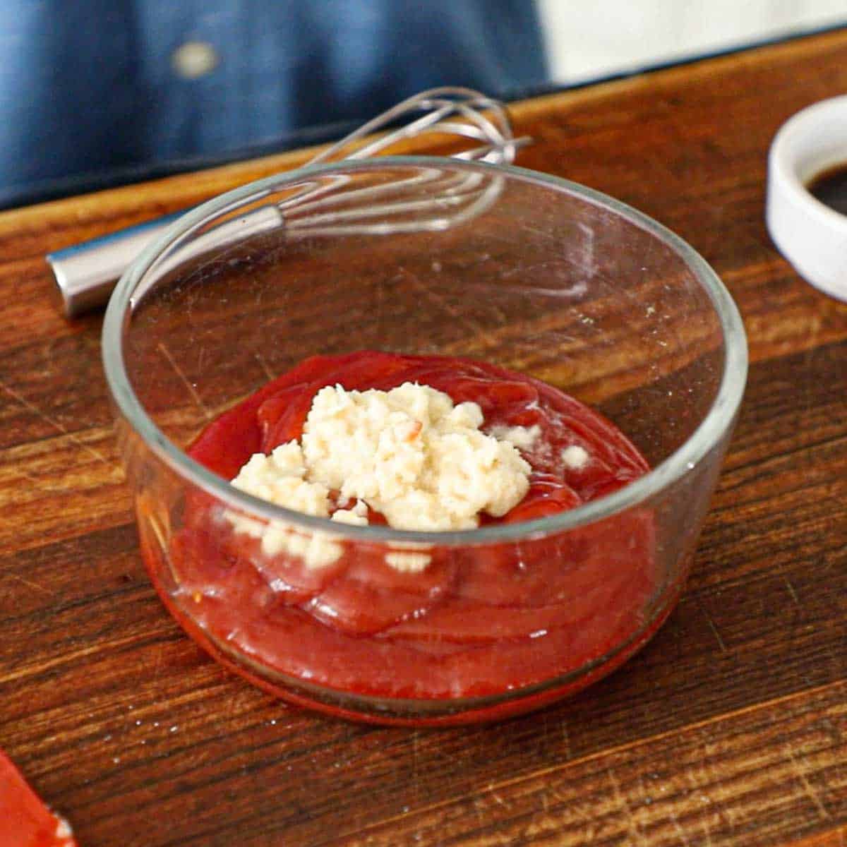 A person standing behind a cutting board that has a glass bowl filled with ketchup and a small pile horseradish sauce on top.