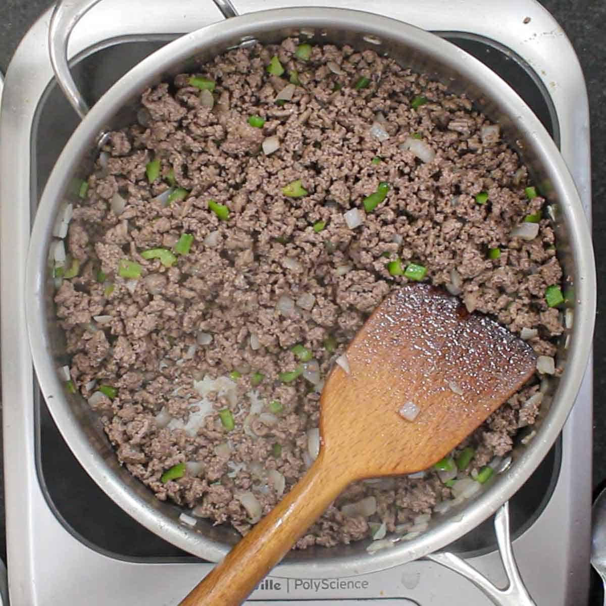 A person using a wooden spatula to stir cooked ground beef along with sautéed onions and green bell pepper in a large silver skillet.