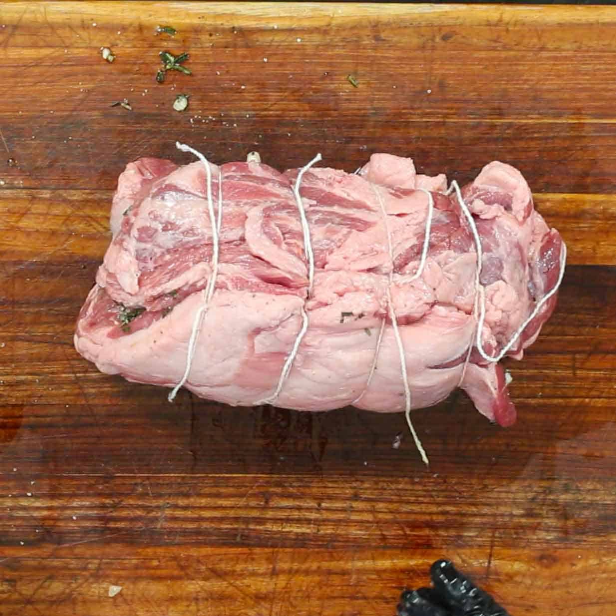 An overhead view of a boneless lamb shoulder that has been rolled and tied together with twine on a wooden cutting board.