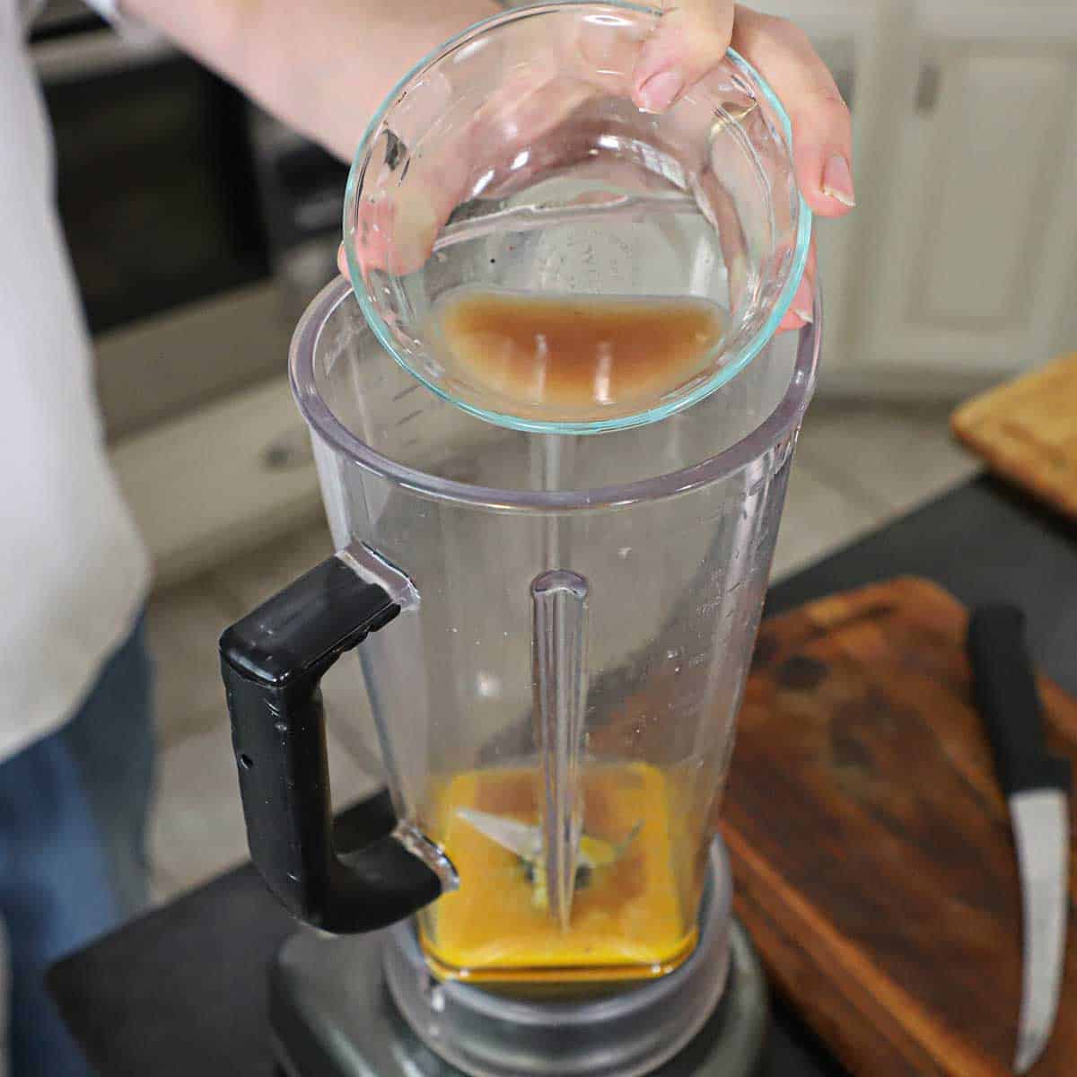 A person pouring a shallot and wine reduction into a blender that contains three egg yolks.