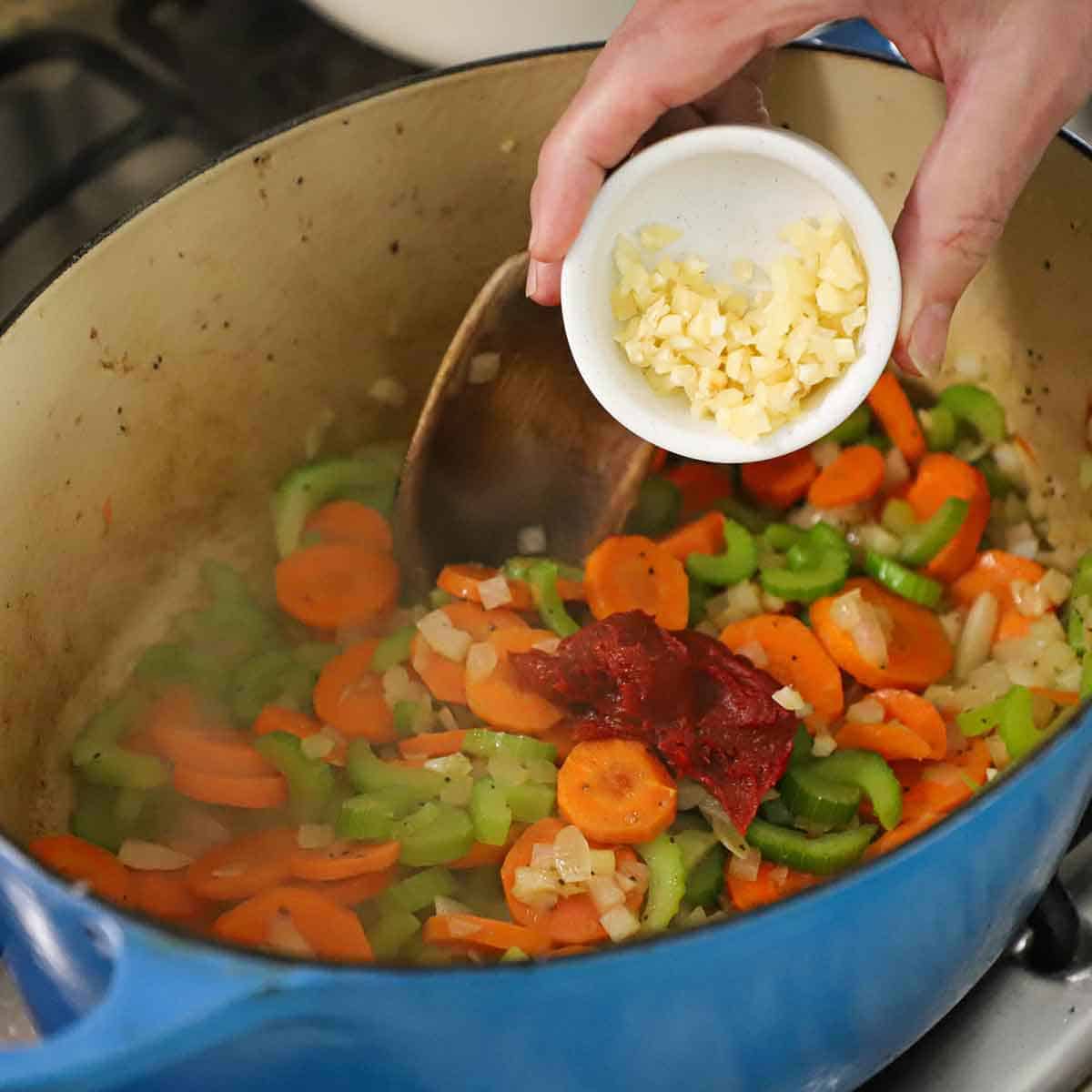 A person dumping minced garlic from a small bowl into a large Dutch oven that is filled with simmering, sautéed carrots, onions, and celery, with a blob of tomato paste sitting on top.