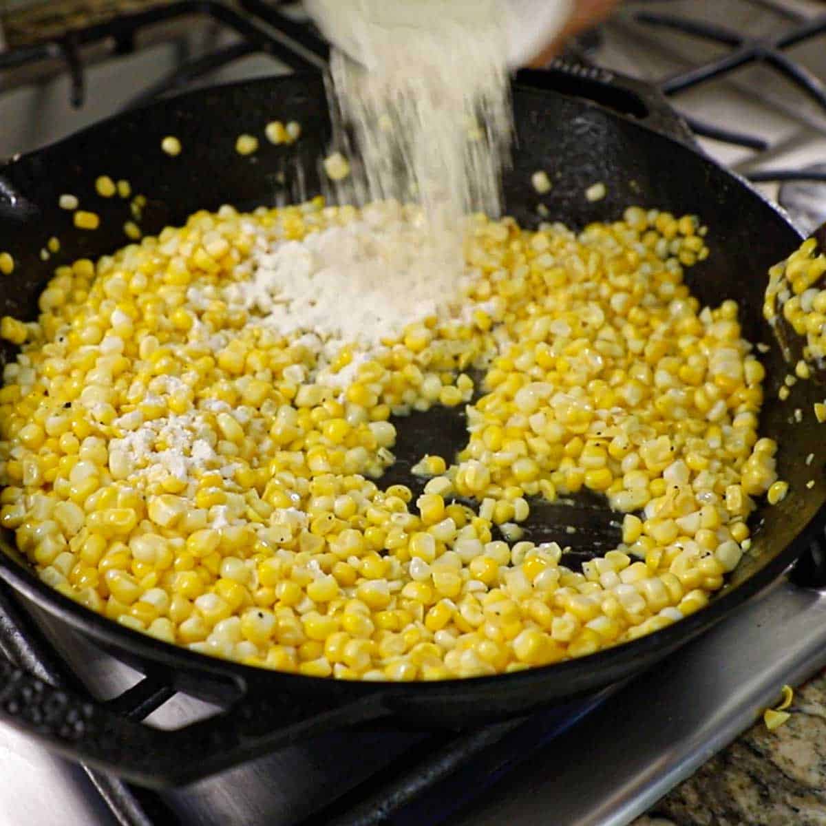 A person adding all-purpose flour from a small bowl over sautéed corn kernels simmering in a large cast-iron skillet on a gas stove.