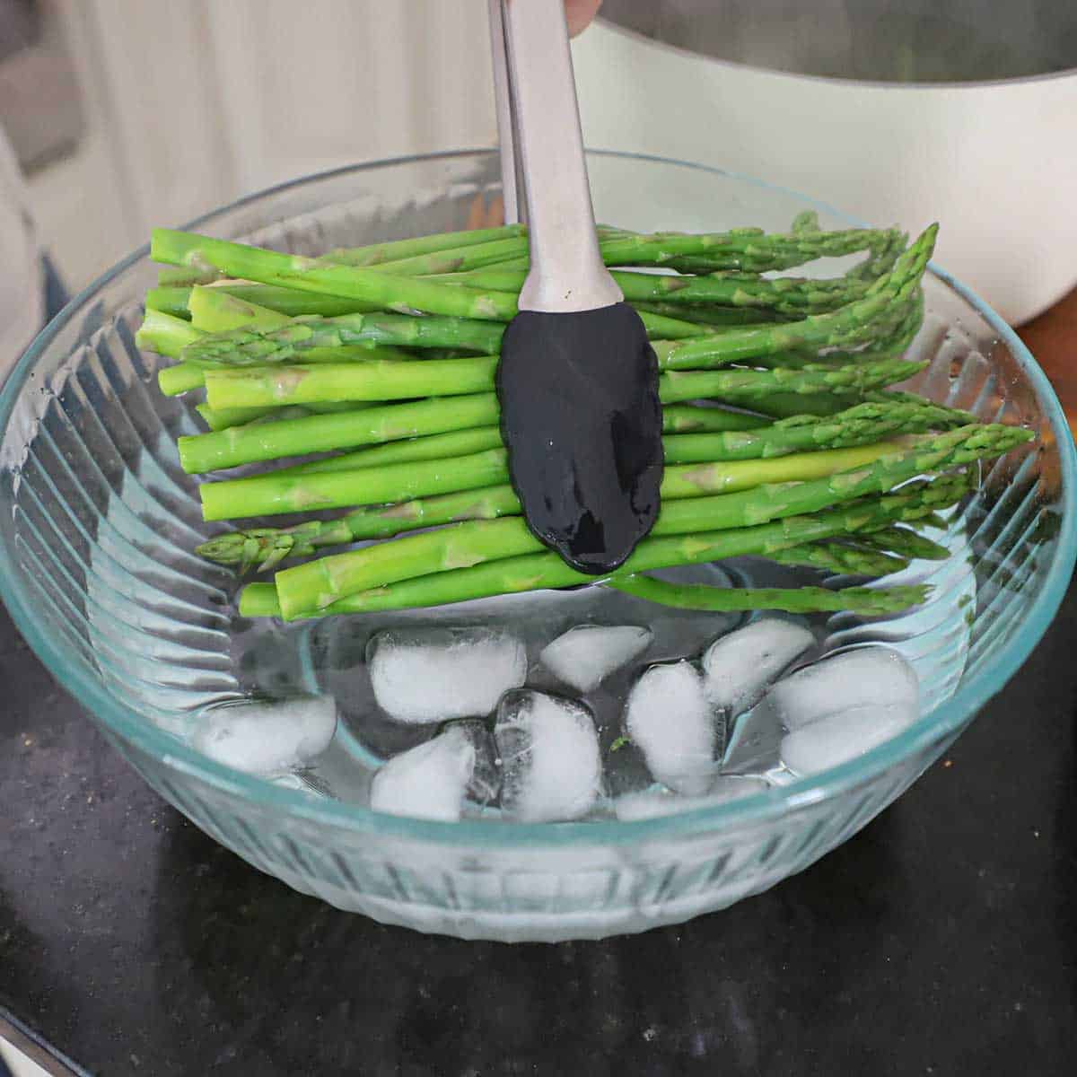 A person adding sprigs of asparagus that have been blanched in boiling water into an ice bath in a large glass bowl.