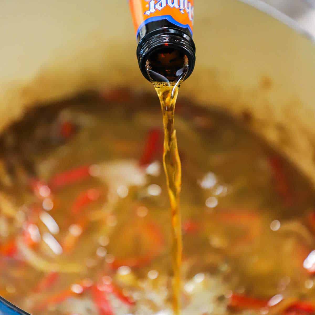 Beer being poured from a beef bottle into a Dutch oven that is filled with a braising liquid for peppers and onions.