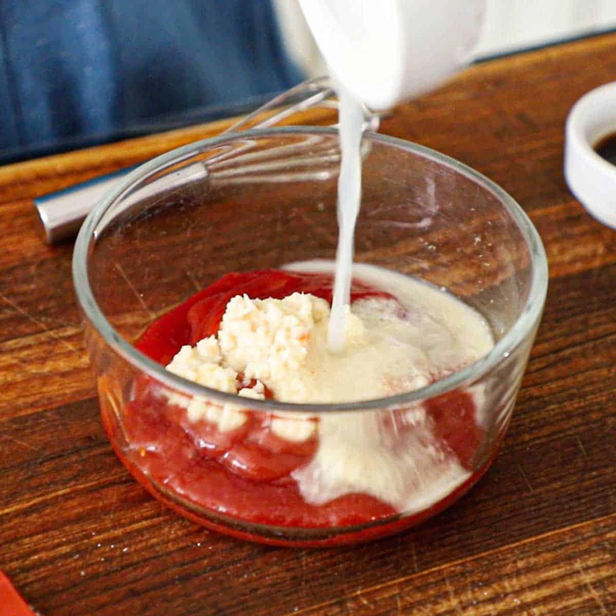 A person pouring lemon juice into a glass bowl filled with ketchup and horseradish sauce on a wooden cutting board.