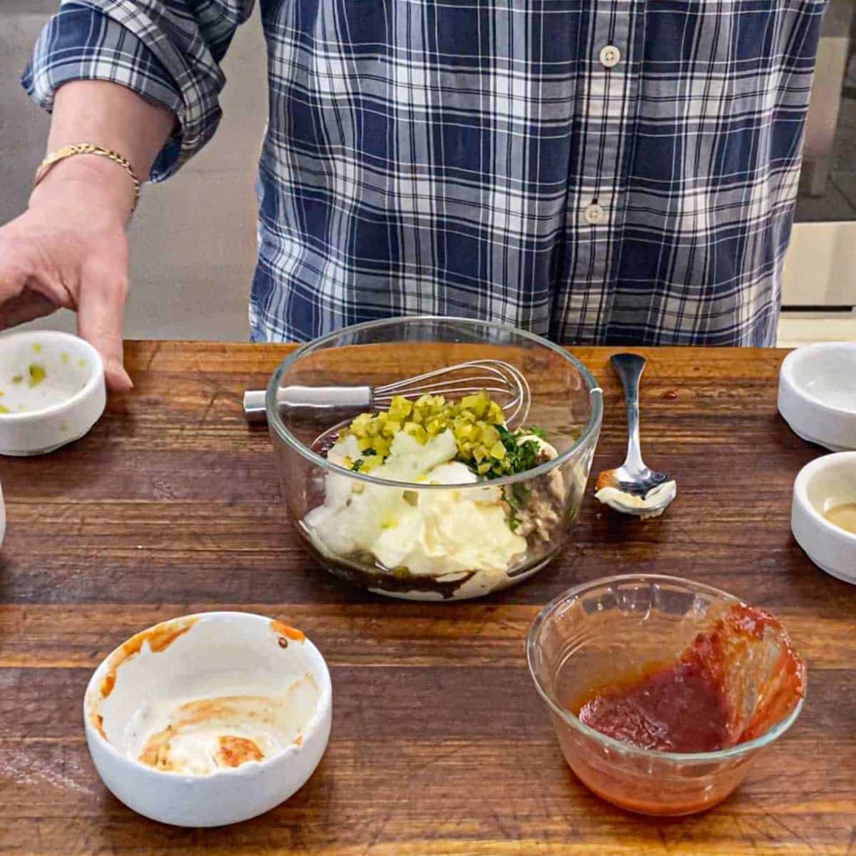 A person holding a small bowl that he just used to dump chopped dill pickles, along with grated onion, and fresh parsley into a bowl with Russian ingredients in it.