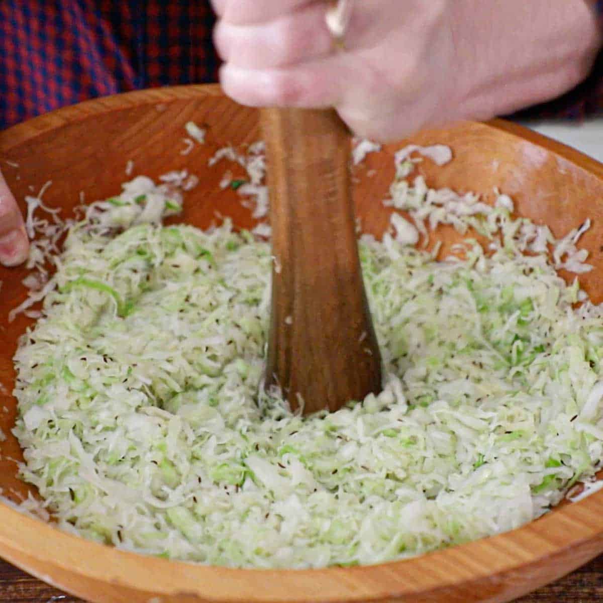 A person using a large wooden rod to crush green cabbage that has been sprinkled with salt in a large wooden bowl.