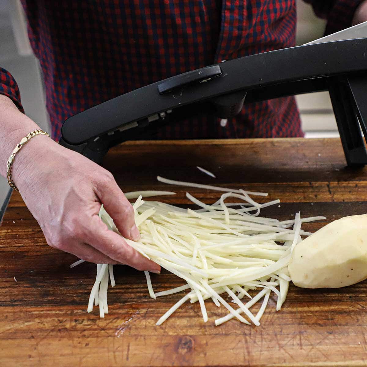 A person lifting freshly julienned shoestring potatoes over a mandolin on a wooden cutting board.