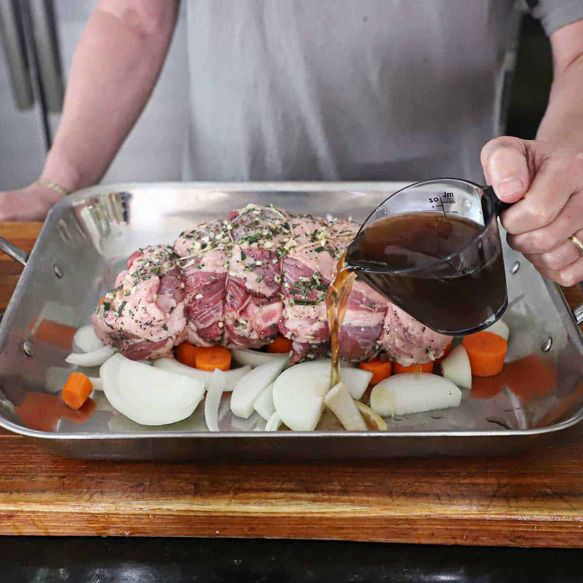 A person pouring beef stock from a measuring cup into a roasting pan filled with a lamb shoulder resting on cut onion and carrots.