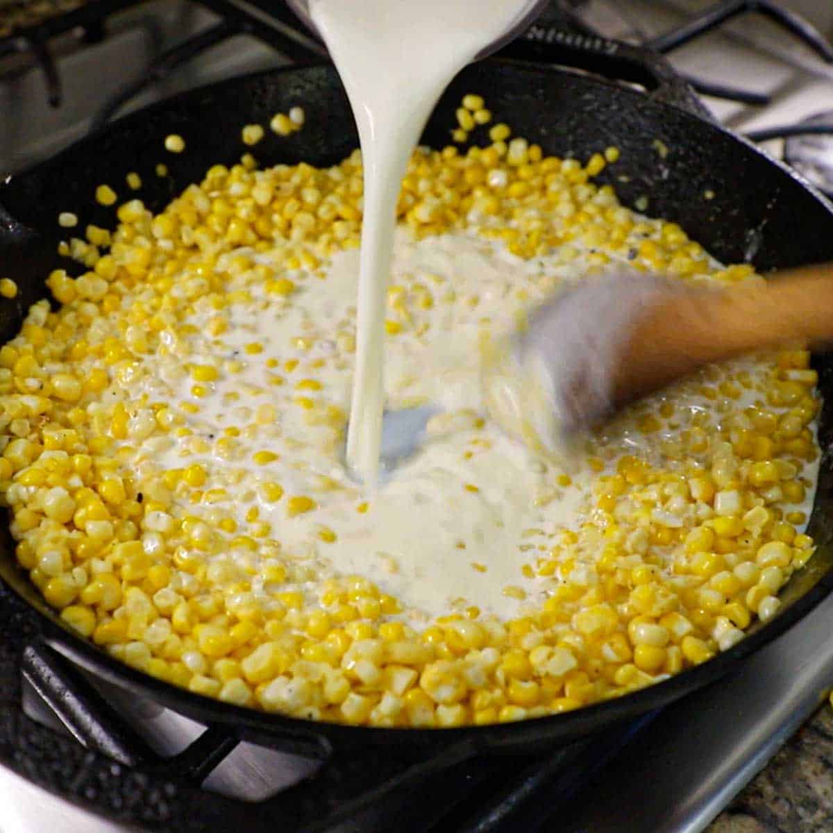 A person pouring cream into a large skillet that is filled with sautéed corn kernels that have been coated in flour.