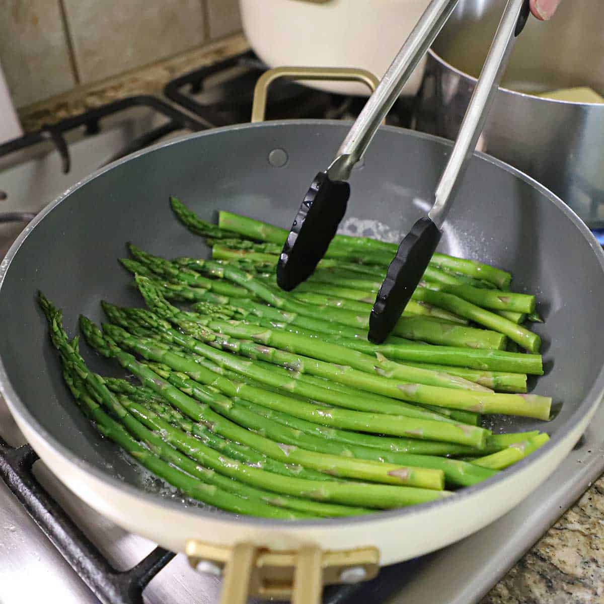 A person using a large pair of tongs to sauté asparagus in a large non-stick skillet with olive oil, butter, salt, and pepper.