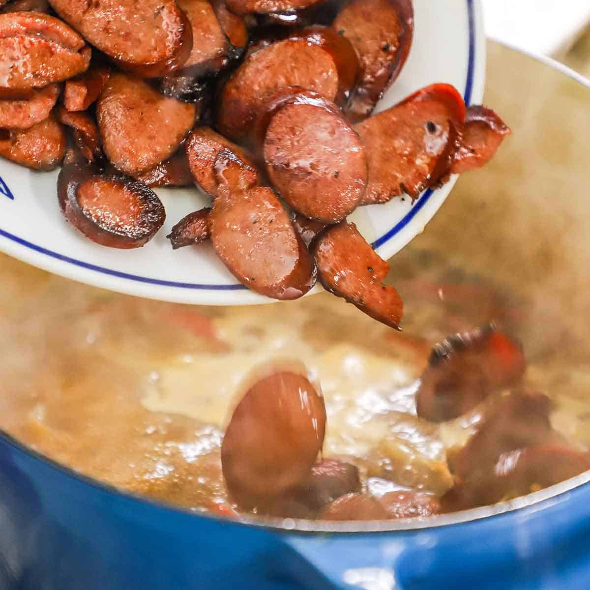 Sautéed sliced kielbasa being transferred from a white platter into a large Dutch oven filled with a simmering liquid with peppers and onions.