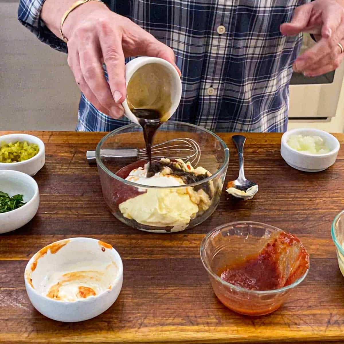 A person pouring Worcestershire sauce into a small glass bowl filled with piles of mayonnaise, chili sauce, sour cream, and horseradish sauce.
