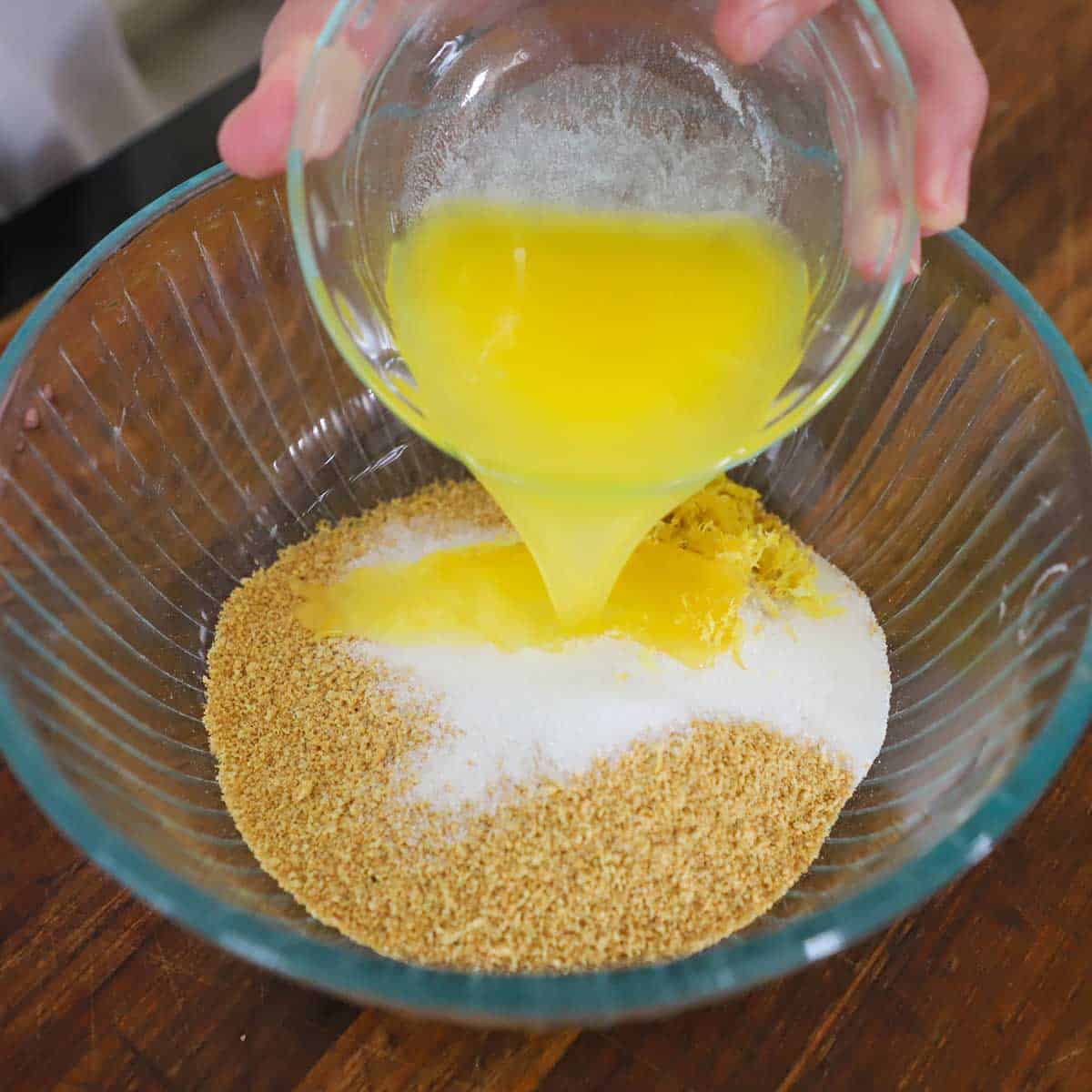 A person pouring melted butter from a small glass bowl into a larger glass bowl filled with graham cracker crumbs and sugar.