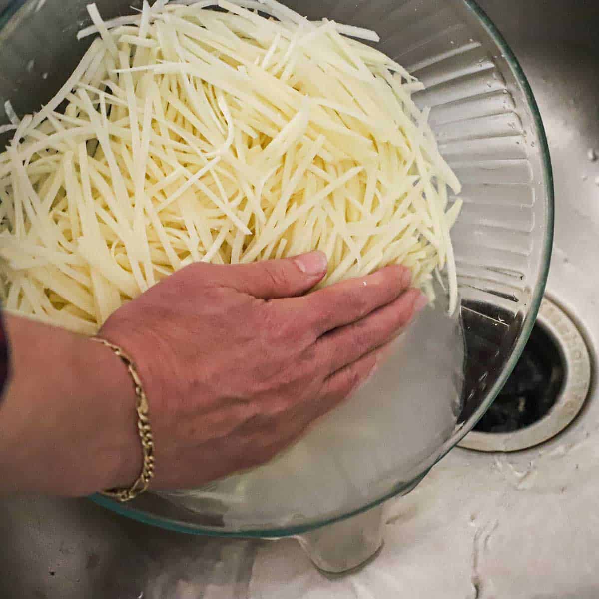 A person using his hand to hold back uncooked shoestring potatoes in a glass bowl with water draining from the bowl into a sink.