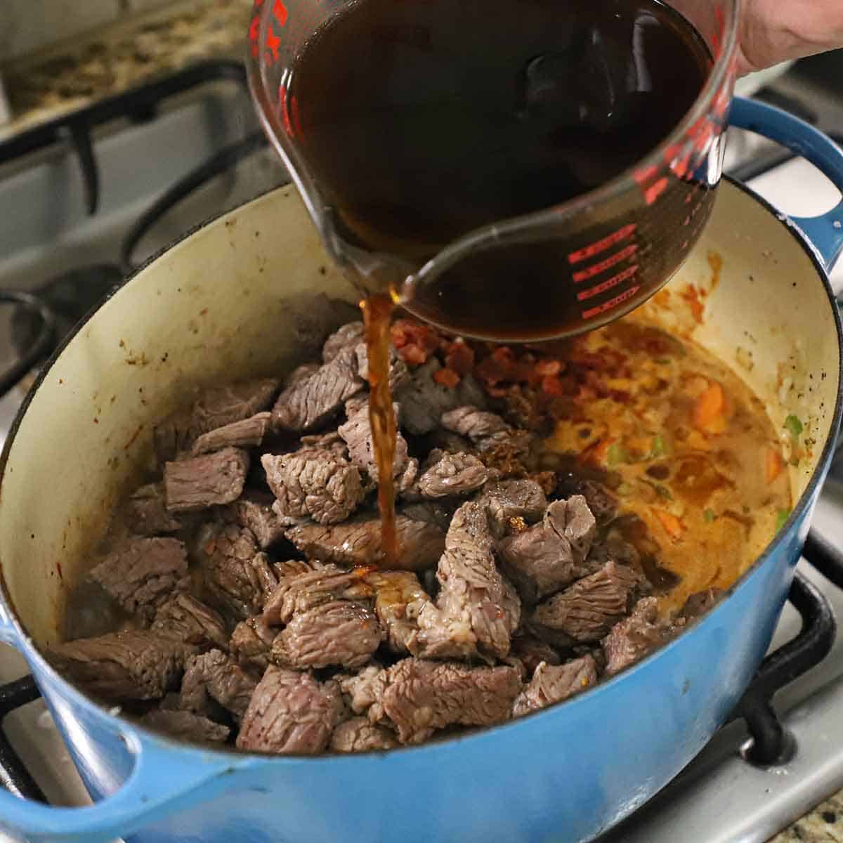 A person pouring four cups of beef broth from a large glass measuring cup into a Dutch oven that is filled with simmering vegetables in beer and topped with seared chunks of beef chuck roast.