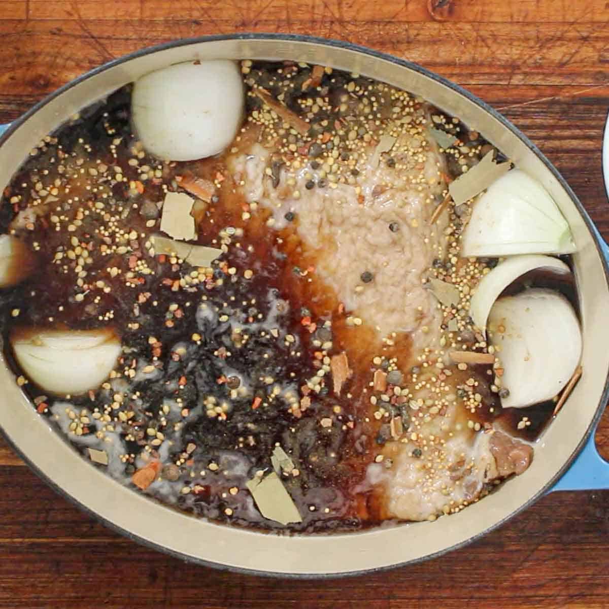 An overhead view of a large oval Dutch oven that is filled with a cured brisket flat surrounded by beef broth, onions, and pickling spices.