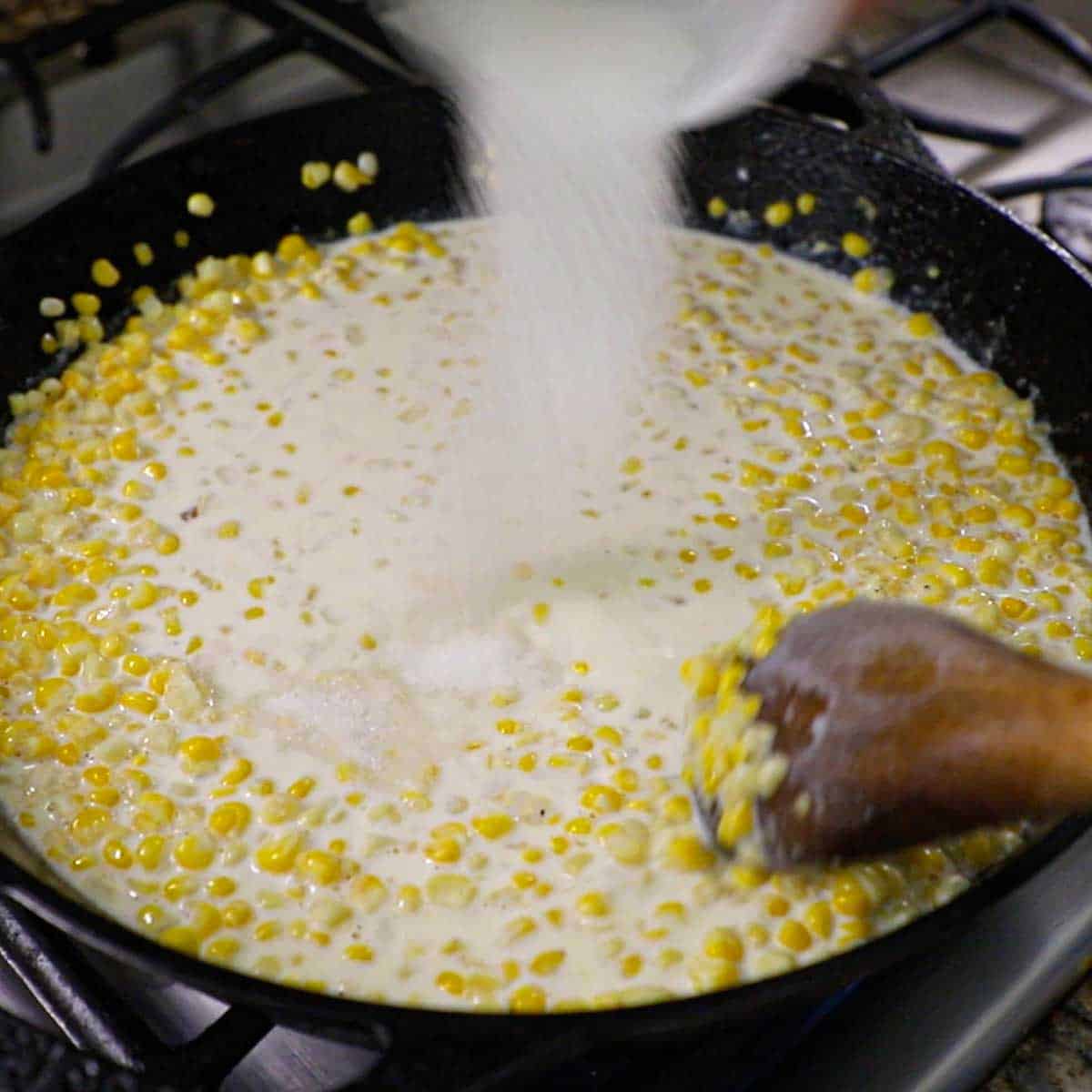 A person transferring sugar from a small bowl into a large cast-iron skillet that is filled with simmering homemade creamed corn.