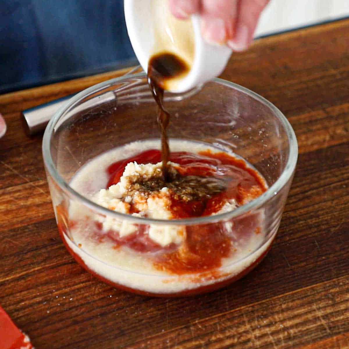 A person pouring Worcestershire sauce from a small white bowl into a larger glass bowl filled with the ingredients of homemade cocktail sauce.