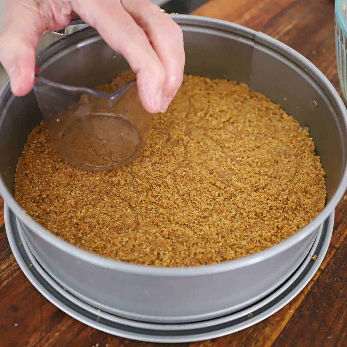 A person using a clear measuring up to compact a graham cracker crust into the bottom of a springform pan.
