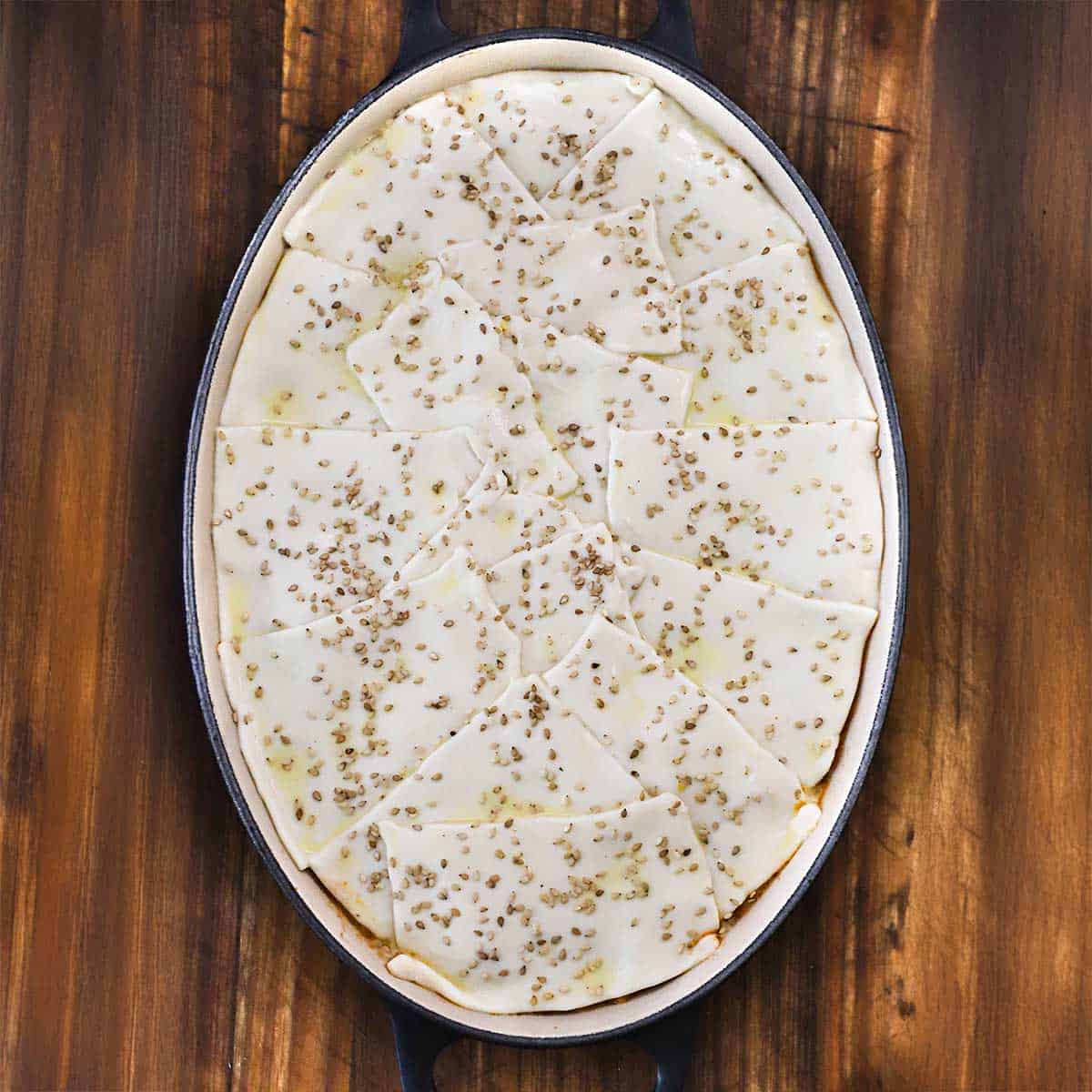 An overhead view of an oval ceramic baking dish that contains an unbaked sloppy Joe casserole topped with pieces of puff pastry, an egg wash, and toasted sesame seeds.