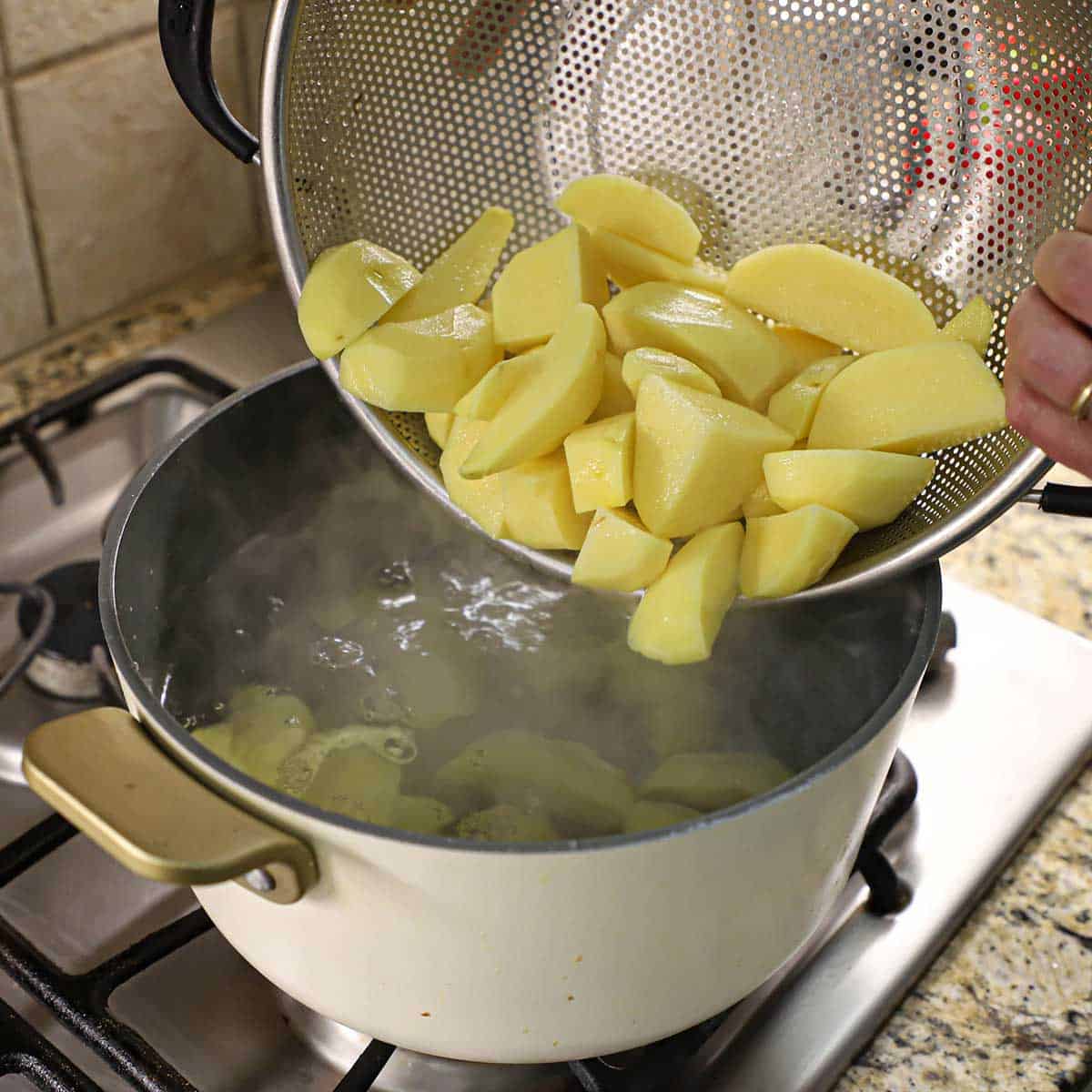 A person transferring gold potato wedges into a pot of boiling water on a gas stove.