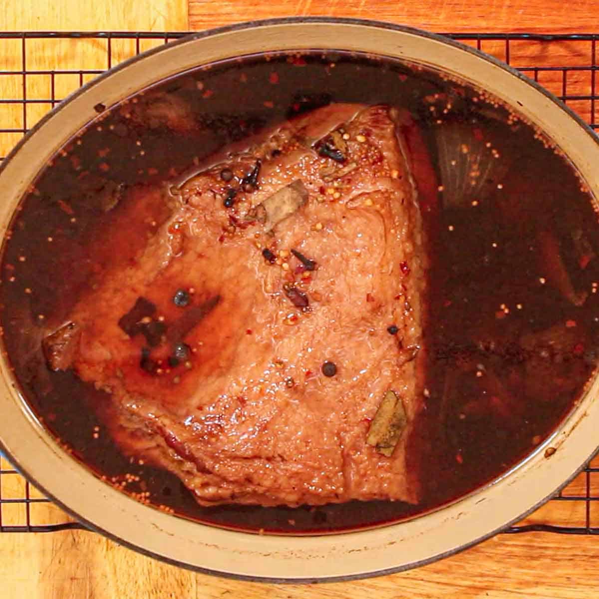 An overhead view of a homemade corned beef brisket that is resting in a braising liquid in a large, oval Dutch oven resting on a baking rack on a wooden cutting board.