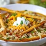A close-up view of a white bowl filled with a serving of white chili with tomatillos and chicken topped with a dollop of sour cream, shredded cheddar, and fried flour tortilla strips.