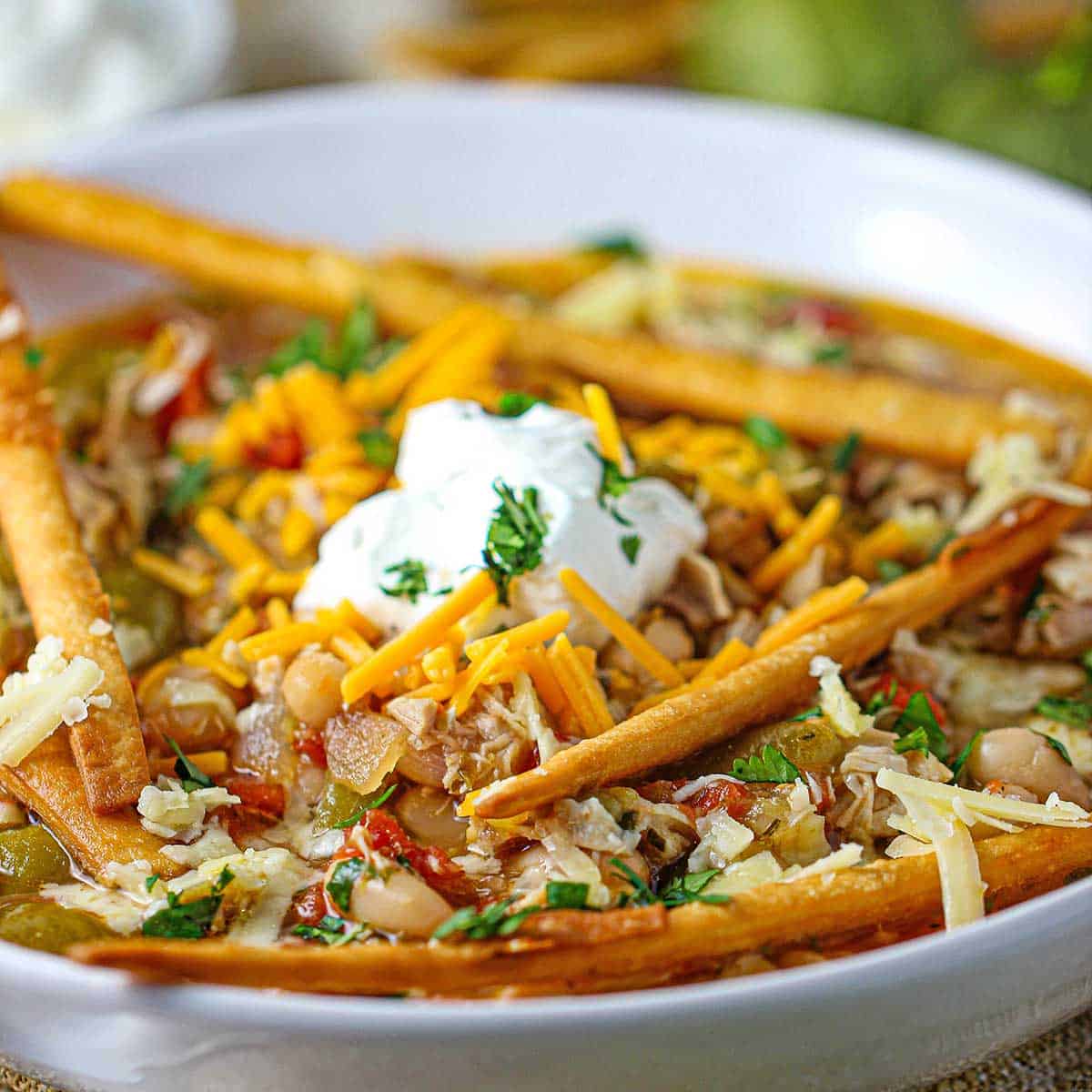 A close-up view of a white bowl filled with a serving of white chili with tomatillos and chicken topped with a dollop of sour cream, shredded cheddar, and fried flour tortilla strips.