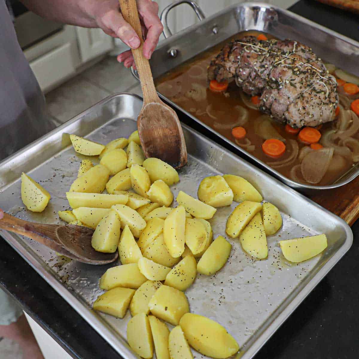 A person using two large wooden spoons to toss par-boiled gold potato wedges in lamb stock and seasonings.
