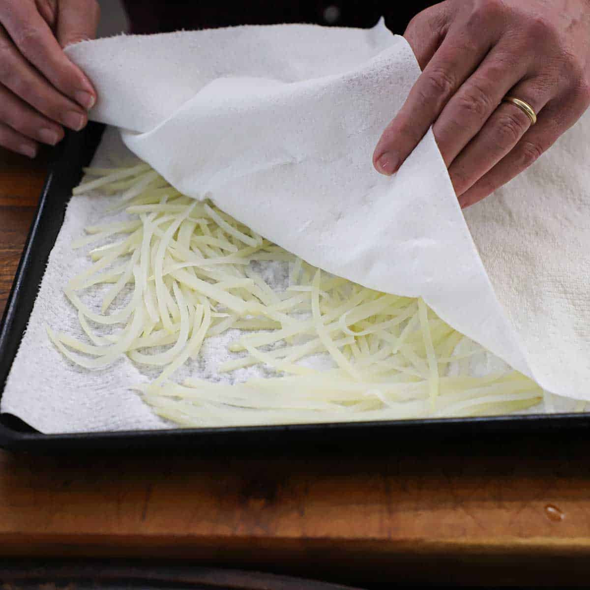 A person holding the corners of paper towels that have are being used to dry wet, uncooked shoestring potatoes that are in a baking pan.