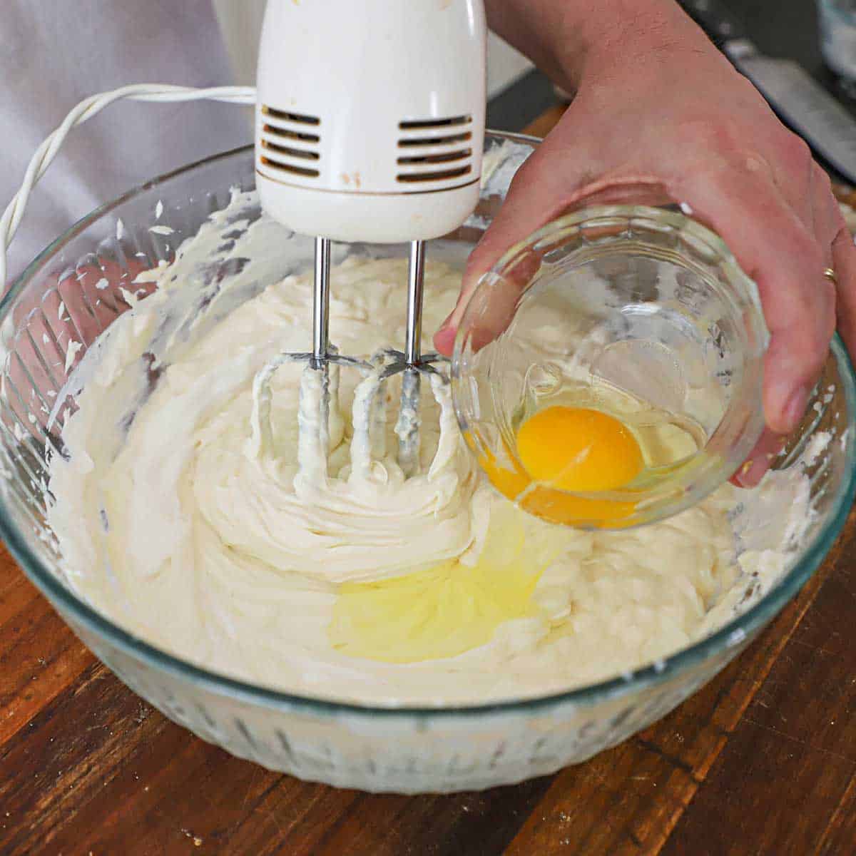 A person transferring an egg into a glass bowl that has a cheesecake filling in along with an electric hand mixer.