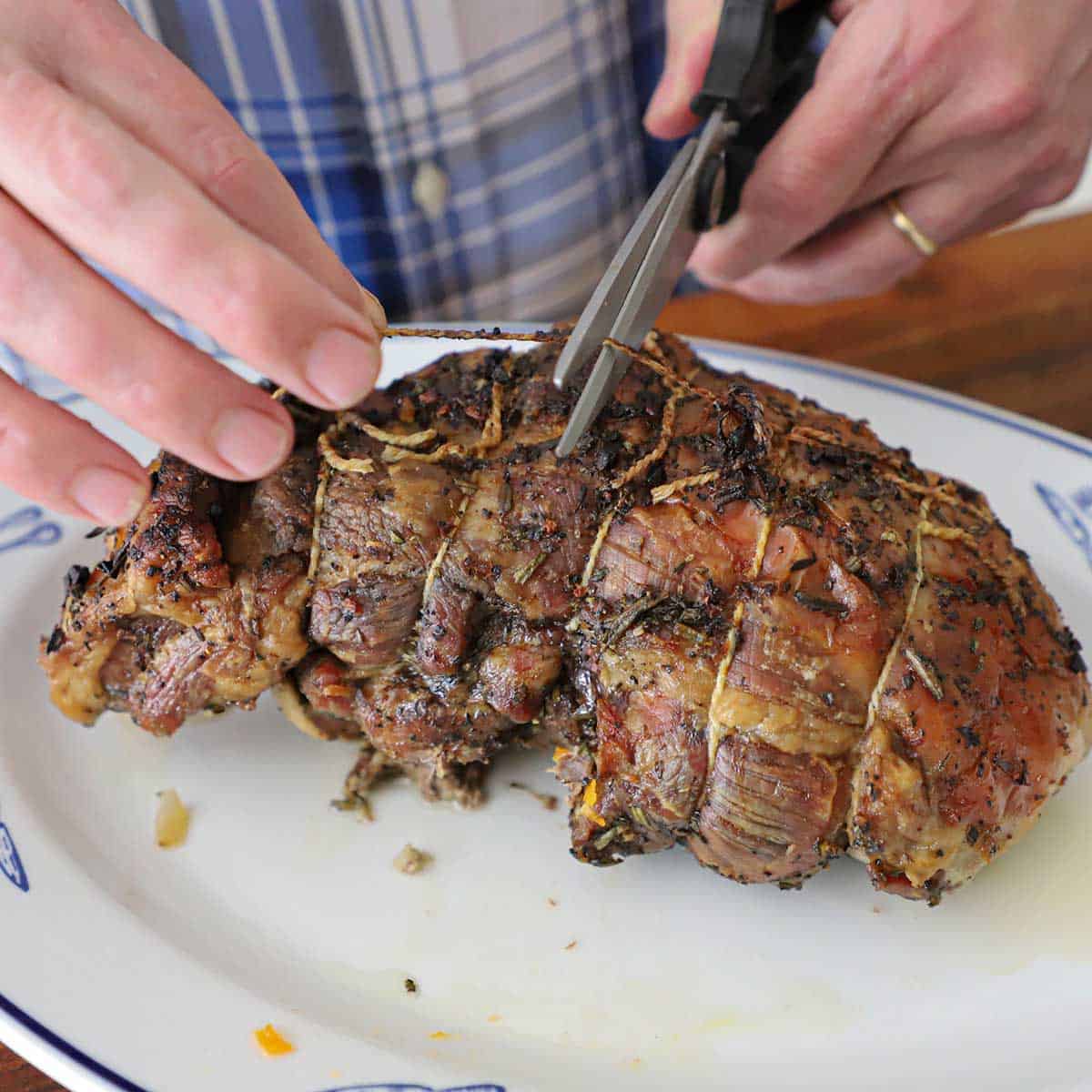 A person using kitchen scissors to cut away twine that is wrapped around a slow-roasted lamb shoulder.